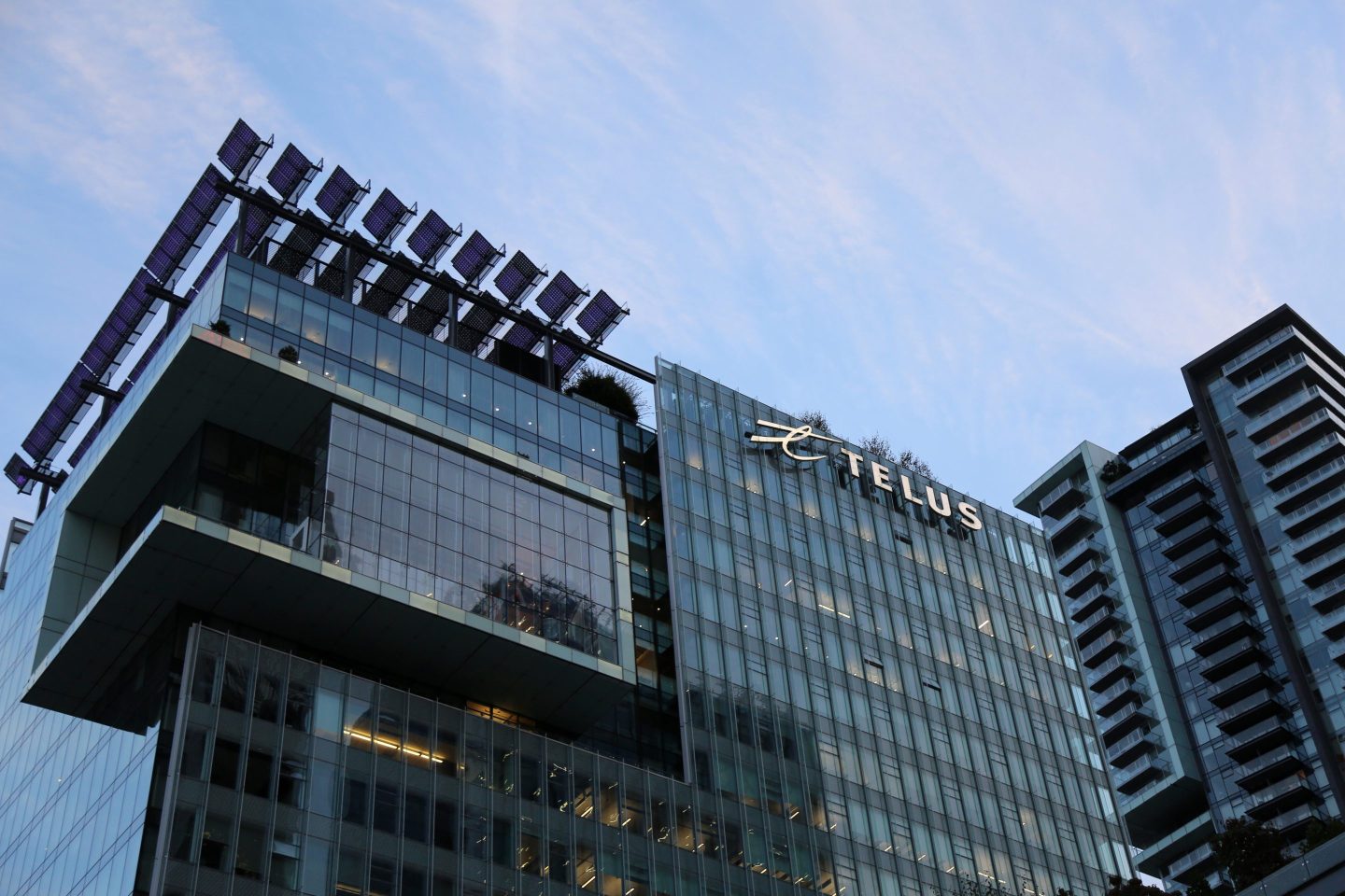 A glass and steel building towers against the blue sky. A sign reading &quot;Telus&quot; is mounted to the side of the building