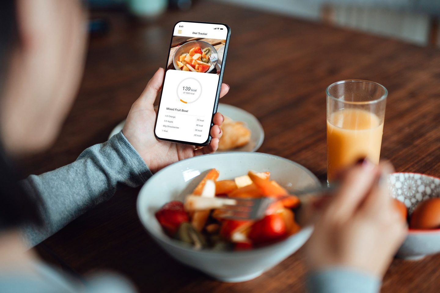 Photo of a young woman using a mobile app to track nutrition and count calories with smartphone while eating breakfast.