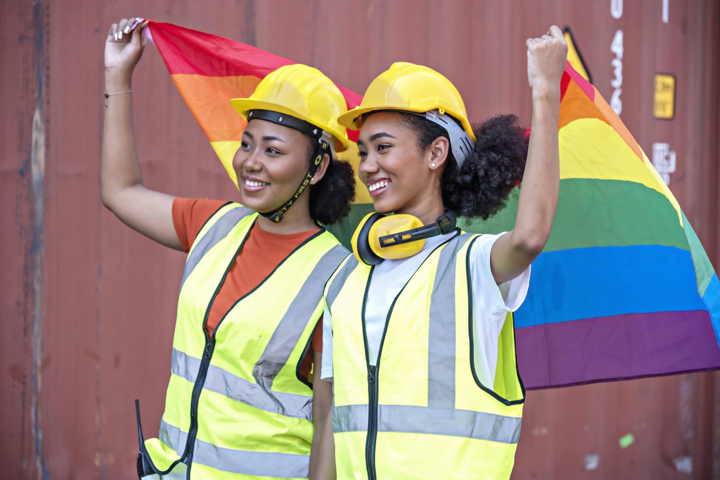 Two women wearing hard hats and reflective vests stand together holding a rainbow LGBTQ pride flag