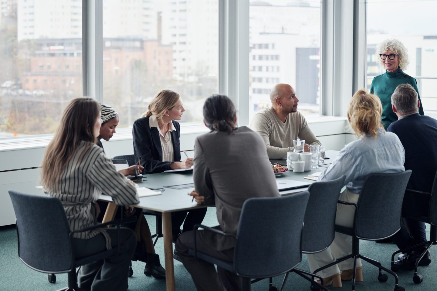 Eight people are gathered around a conference table with windows facing the city skyline