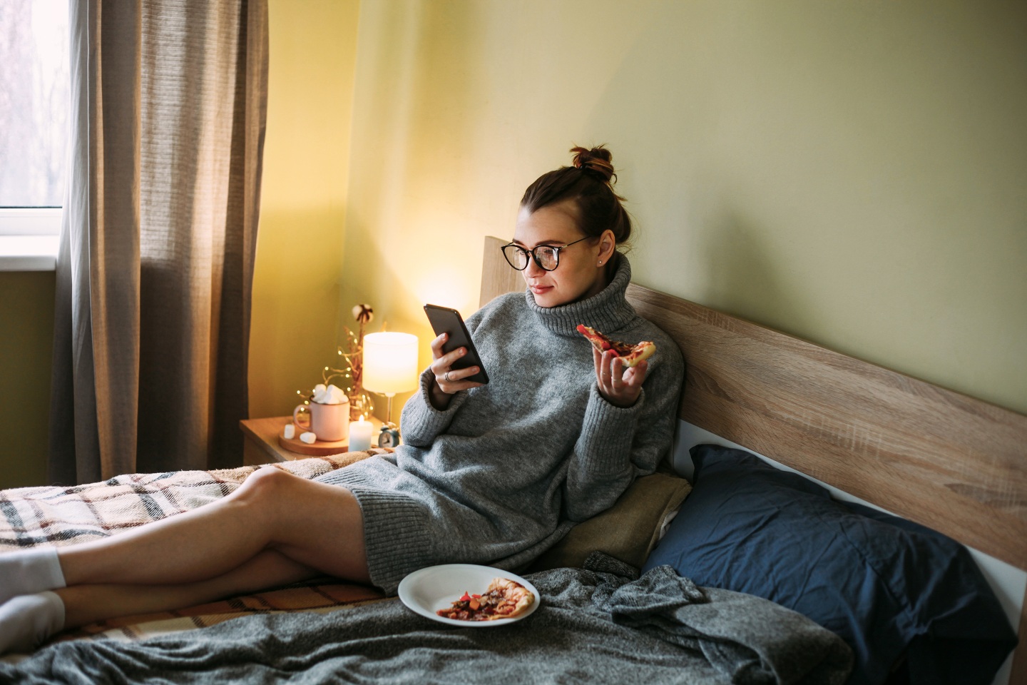 Woman using her phone and eating pizza in bed.